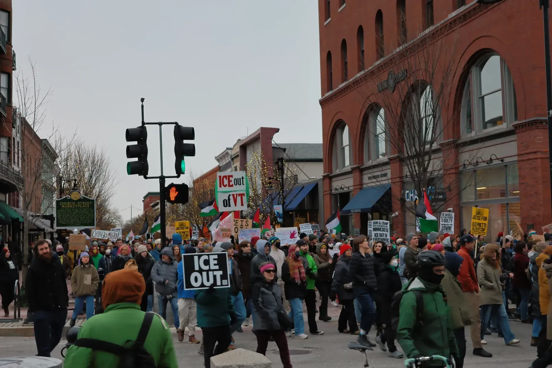 Crowd of protesters marching past a brick building, holding signs including “ICE OUT,” at an intersection.