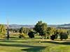 Sunlit golf course with green fairways, trees, and hills under a clear blue sky.