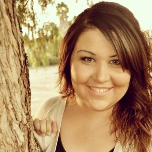 A smiling person standing beside a tree trunk in an outdoor setting with warm, sepia-toned lighting.