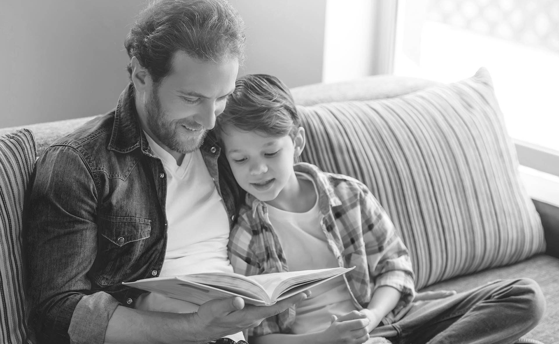 Two people reading a book together on a couch, smiling in a bright room.