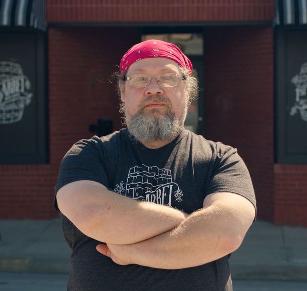 A man with a beard and glasses is standing with his arms crossed in front of a brick building.