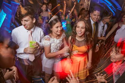 People dancing at a party; girl in a white dress smiles between two friends. Blurred lights and movement.