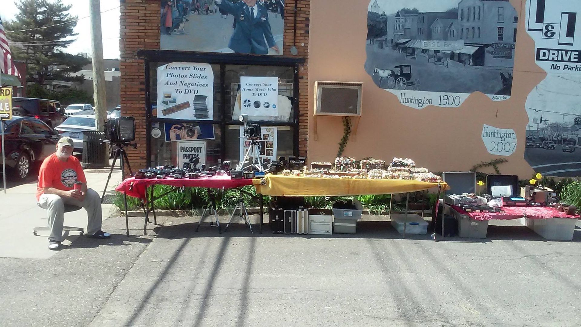 Outdoor craft fair display with tables of colorful items beside a storefront, with a person seated at left.