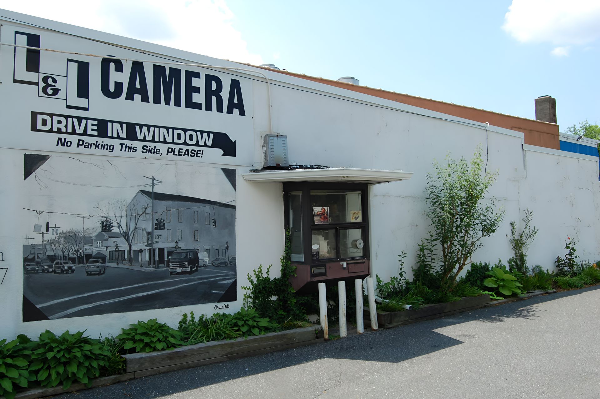 L Camera storefront with large mural and drive-in window on a sunny street side wall