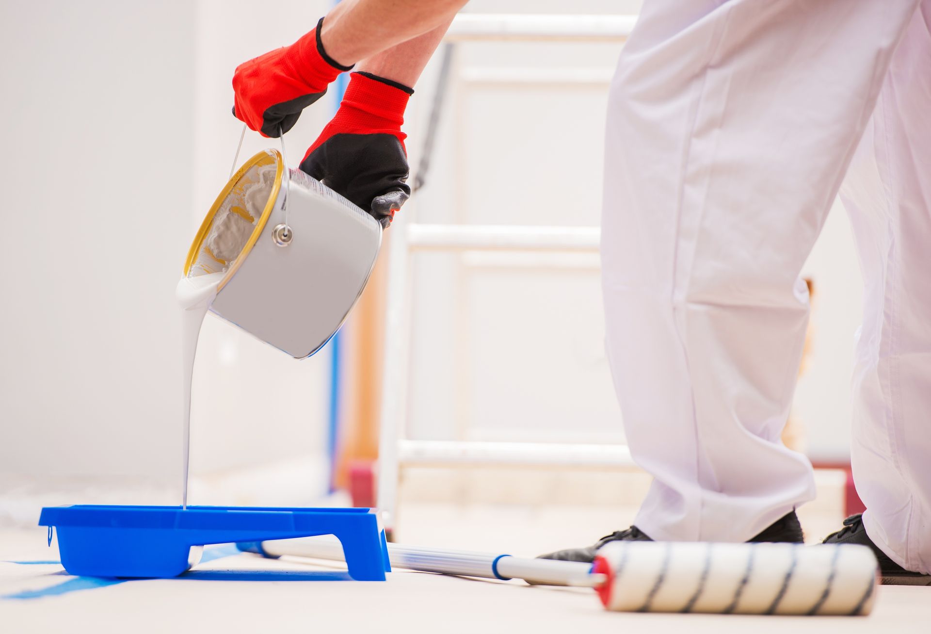 A person wearing white work pants and red-palmed gloves pours white paint from a bucket into a blue paint tray.