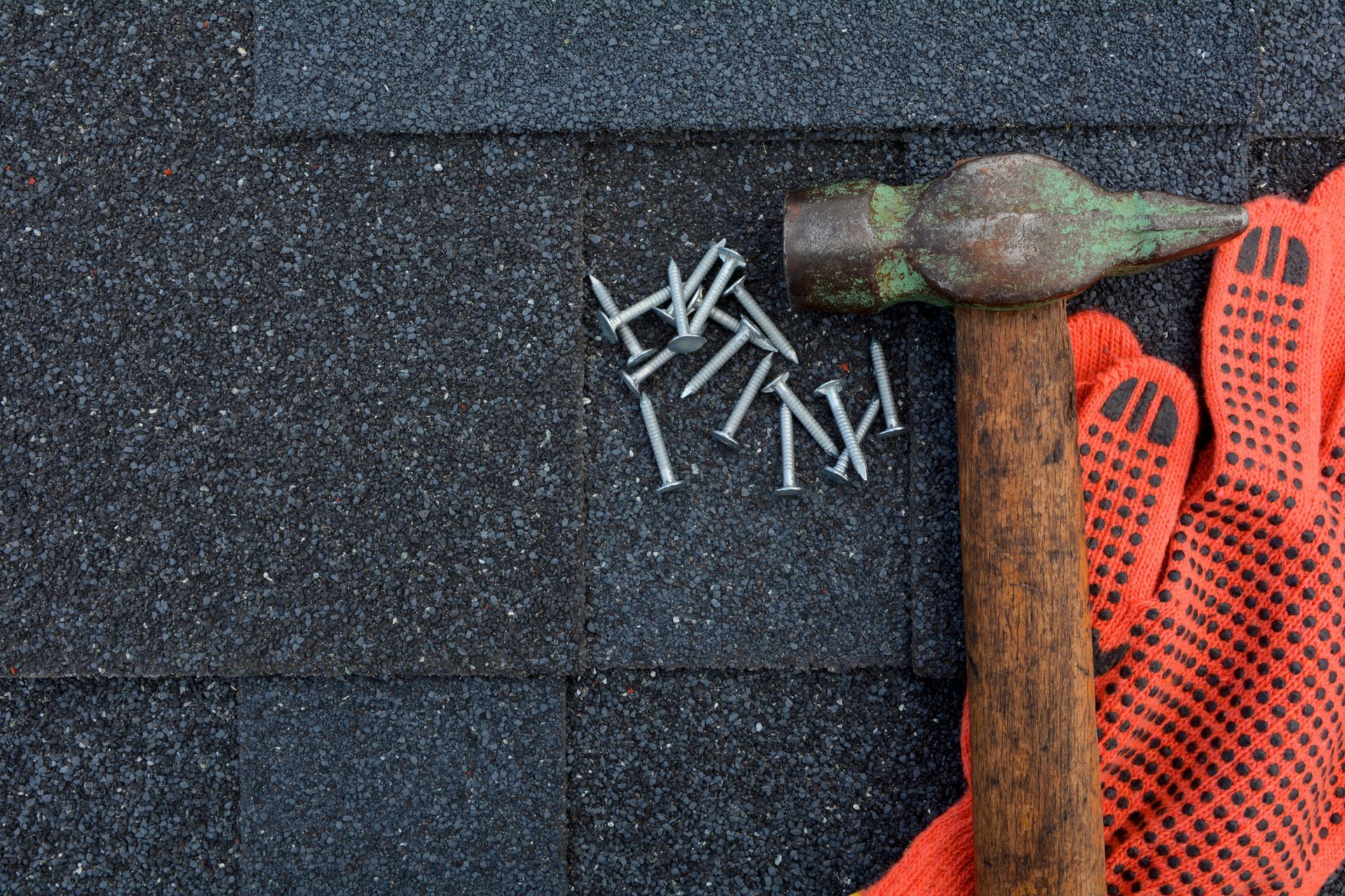 A hammer and a pile of roofing nails lie next to orange work gloves on black asphalt roofing shingles.