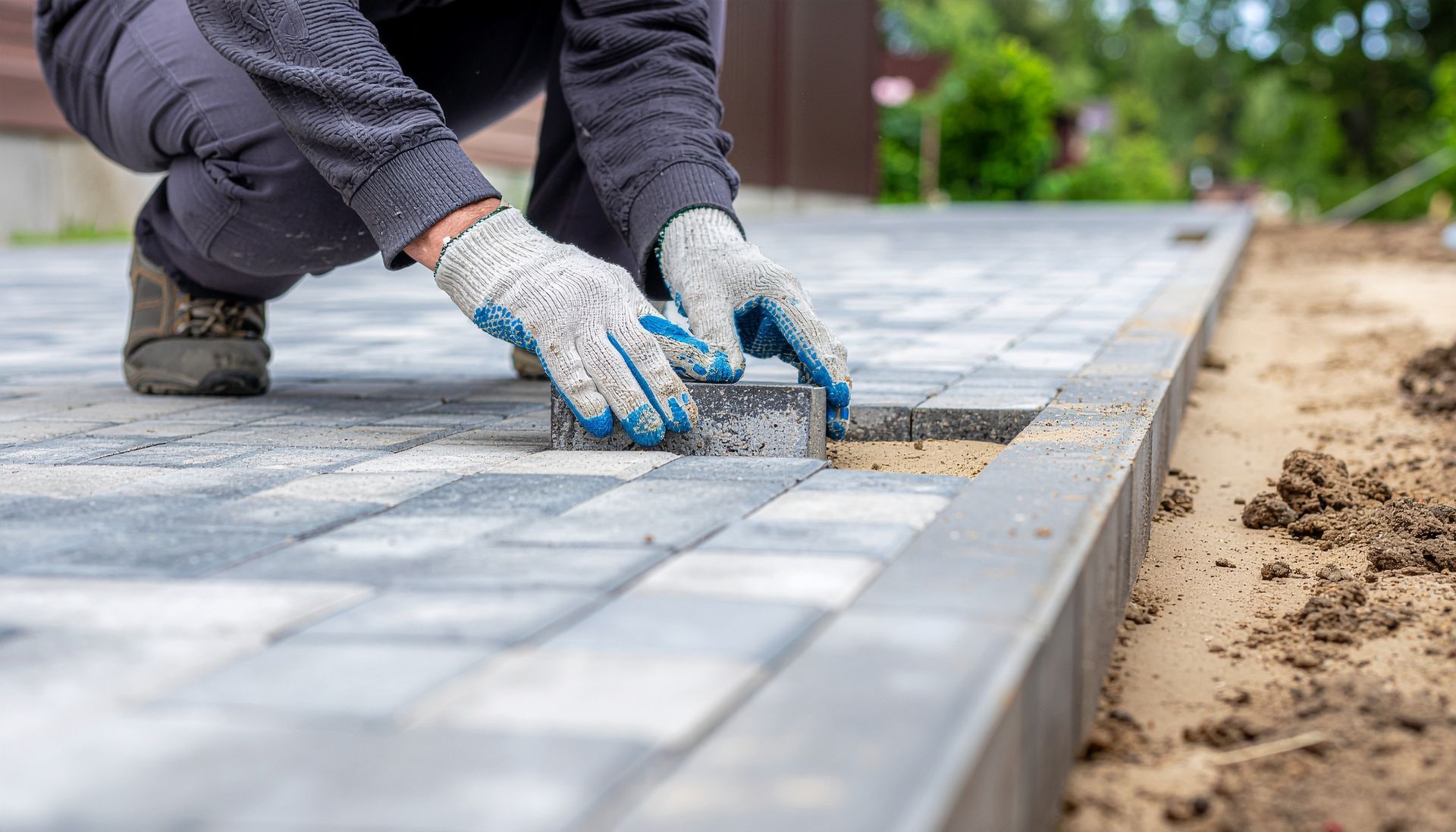 A person wearing work gloves places a gray stone paver into a paved walkway next to a dirt edge.