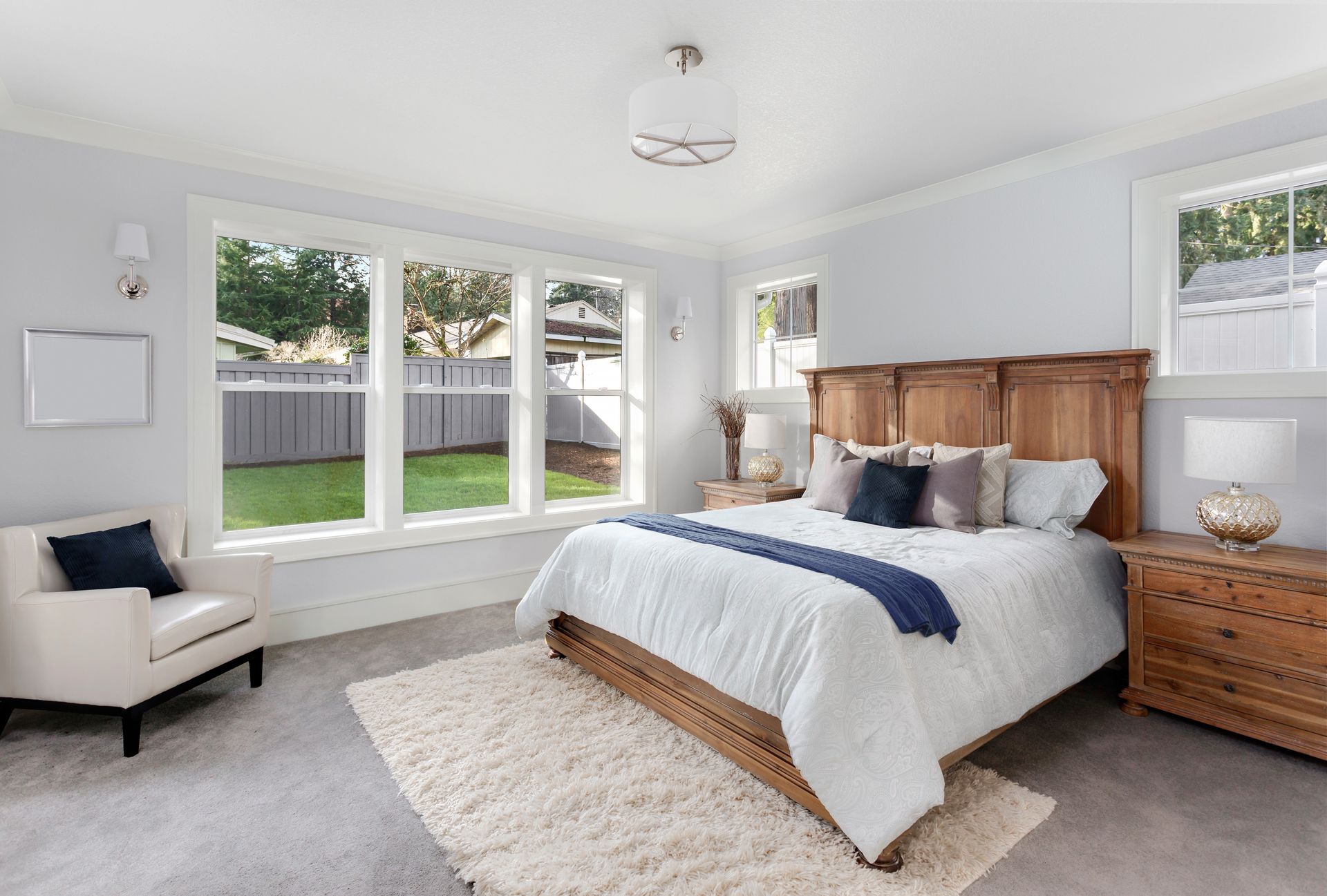 A bright bedroom with a wooden bed, white bedding, a cream armchair, and a rug against a backdrop of large windows. A bright bedroom with a wooden bed, white bedding, a cream armchair, and a rug against a backdrop of large windows.
