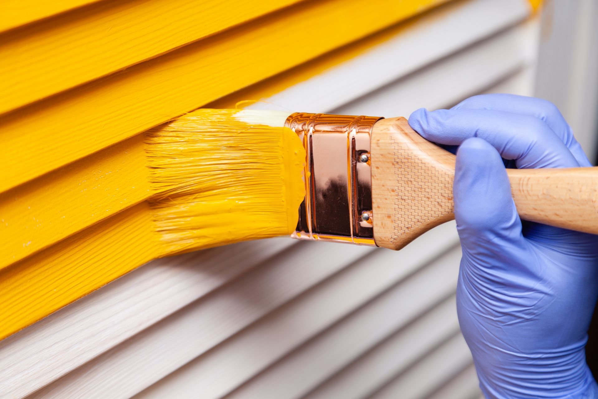 A hand wearing a blue nitrile glove uses a brush to paint white louvered blinds with bright yellow paint. A hand wearing a blue nitrile glove uses a brush to paint white louvered blinds with bright yellow paint.