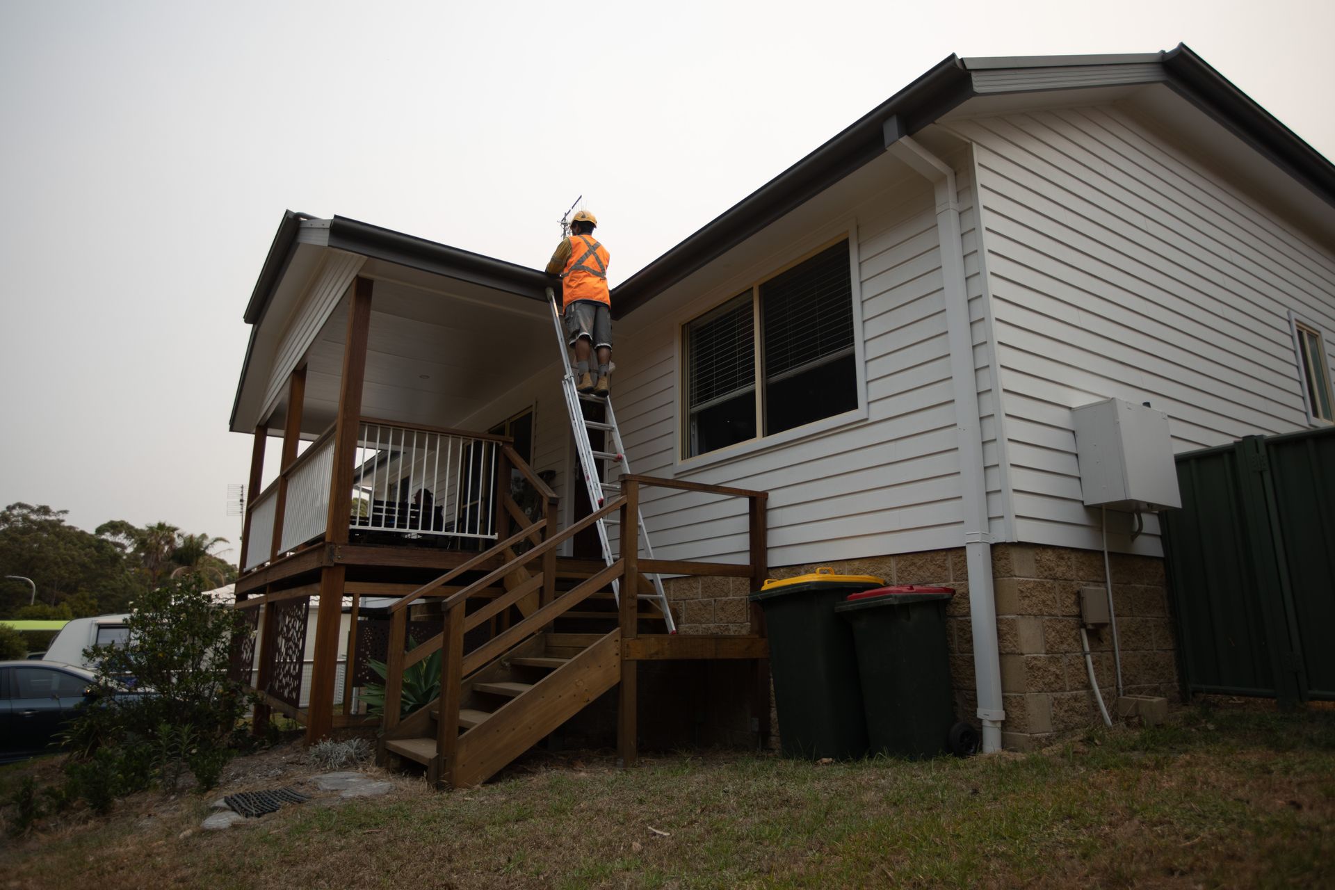 A person in an orange high-visibility vest climbs a ladder against the side of a white house to inspect the roof. A person in an orange high-visibility vest climbs a ladder against the side of a white house to inspect the roof.
