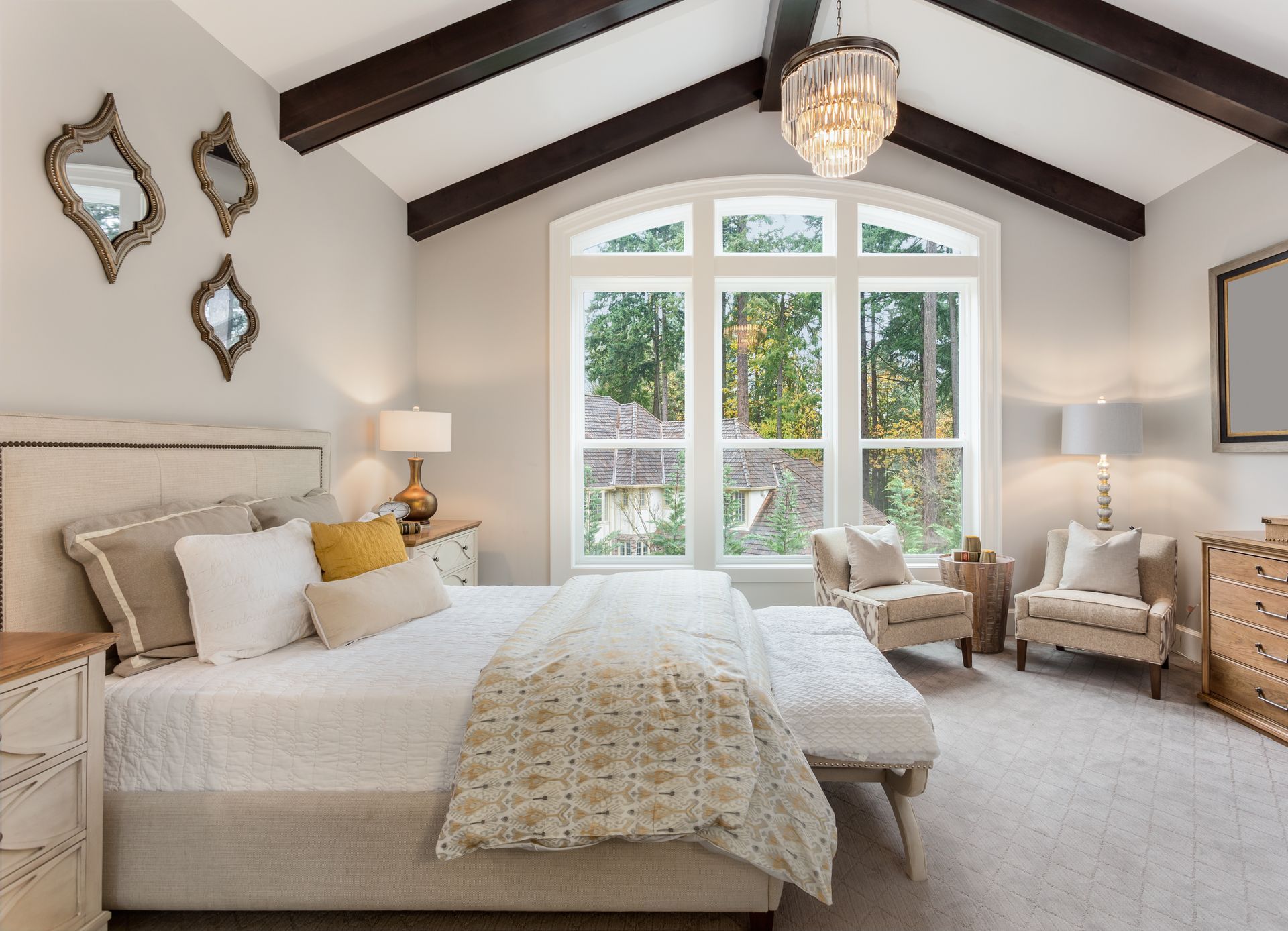 A cozy bedroom featuring a queen bed, vaulted ceiling with dark wooden beams, large windows, and neutral-toned decor.