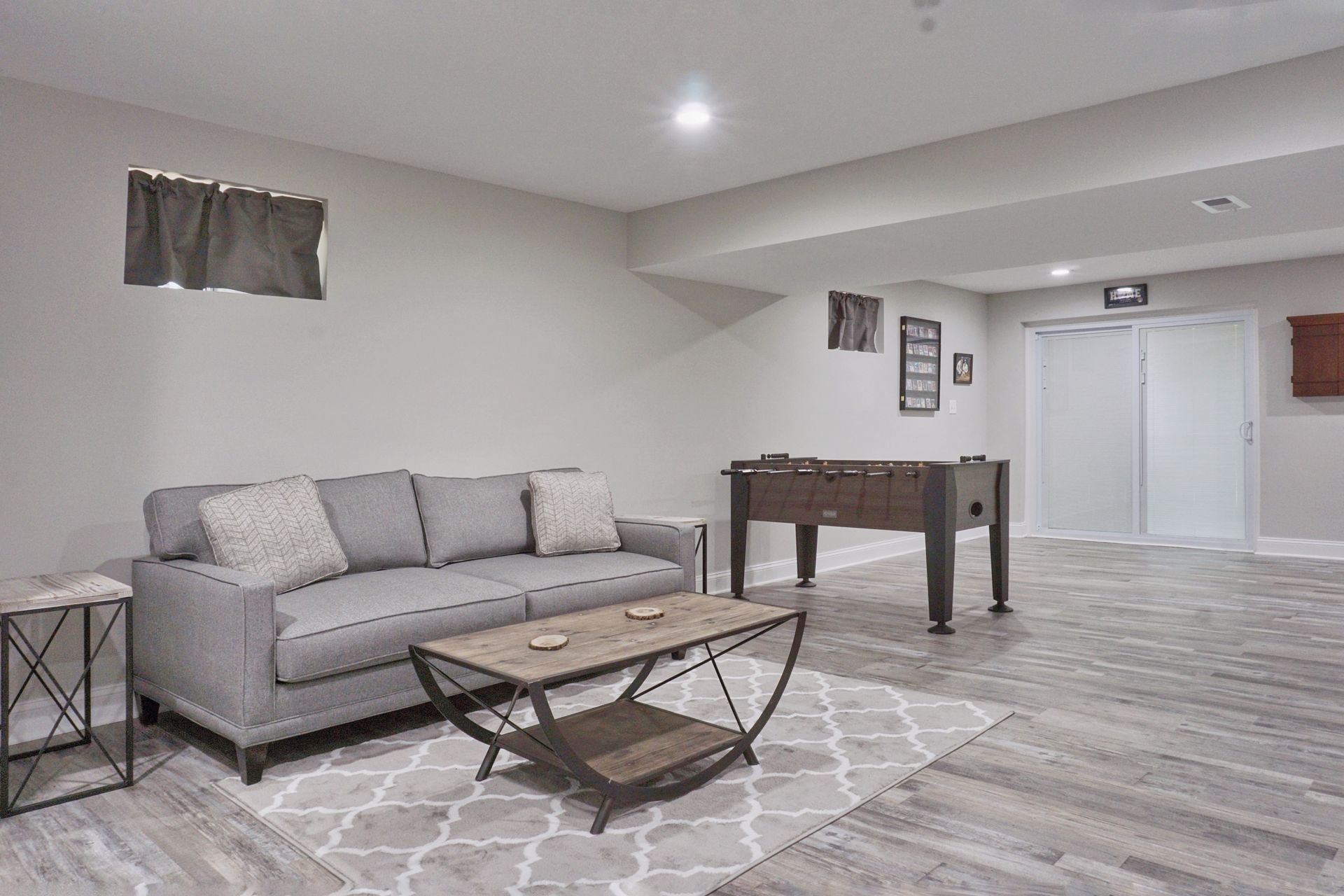 A finished basement featuring a gray sofa on a patterned rug, a wooden coffee table, and a foosball table.