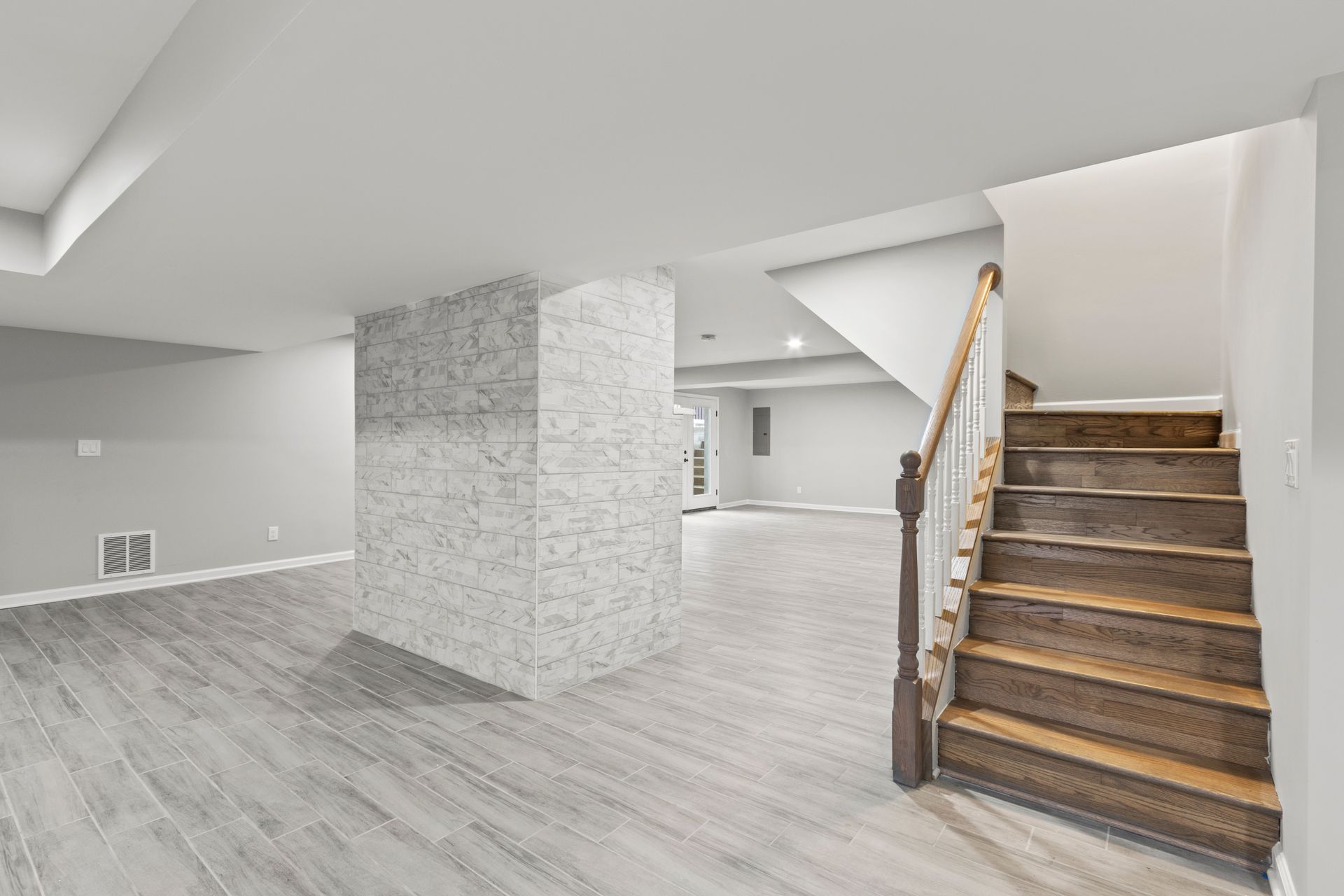A finished basement featuring light gray wood-look flooring, a central stone-clad support pillar, and wooden stairs.