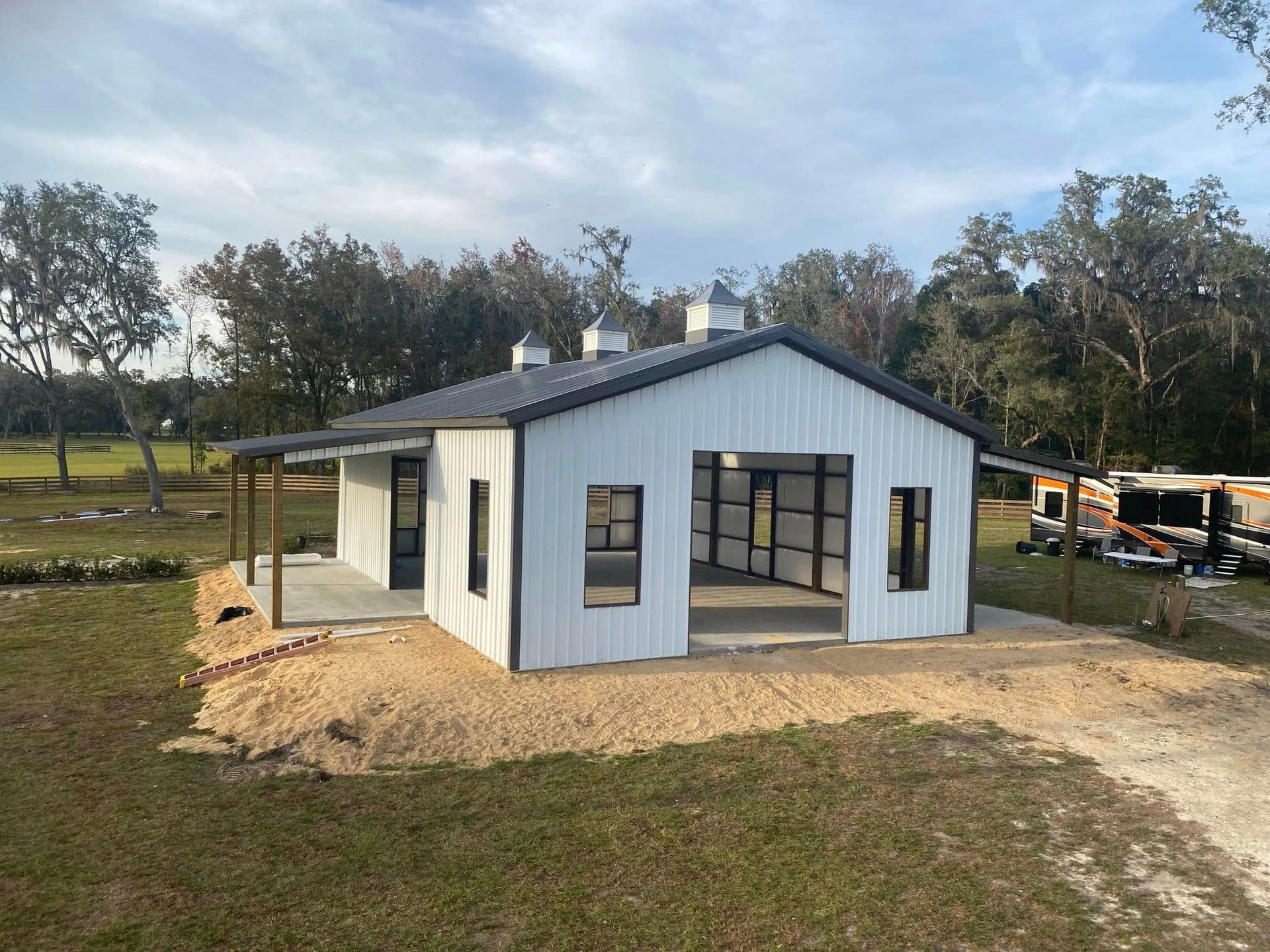 Small white house with a gray roof on a rural lot, with trees and construction materials nearby