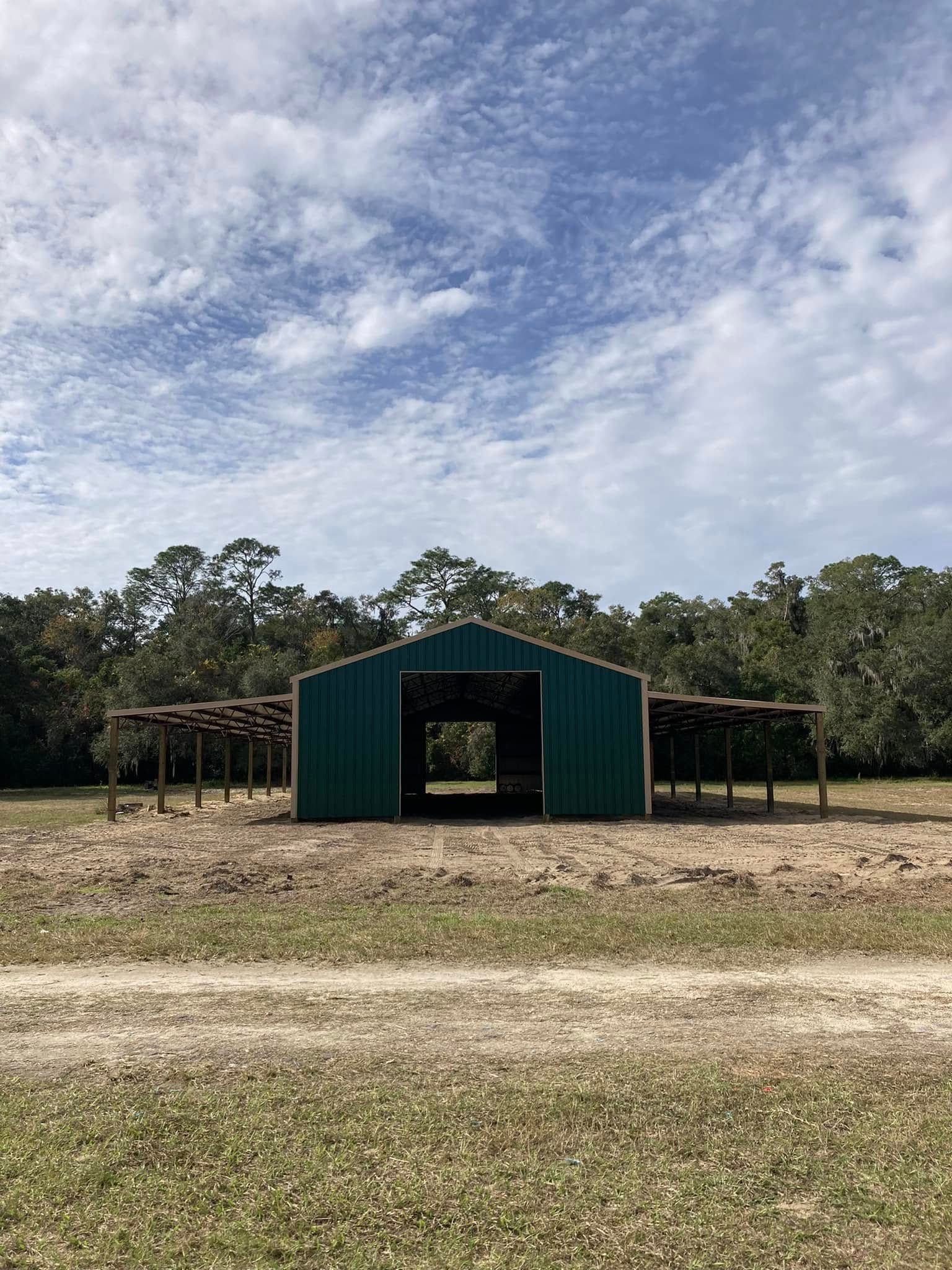 Green metal barn centered in a grassy field under a cloudy sky, with trees in the background