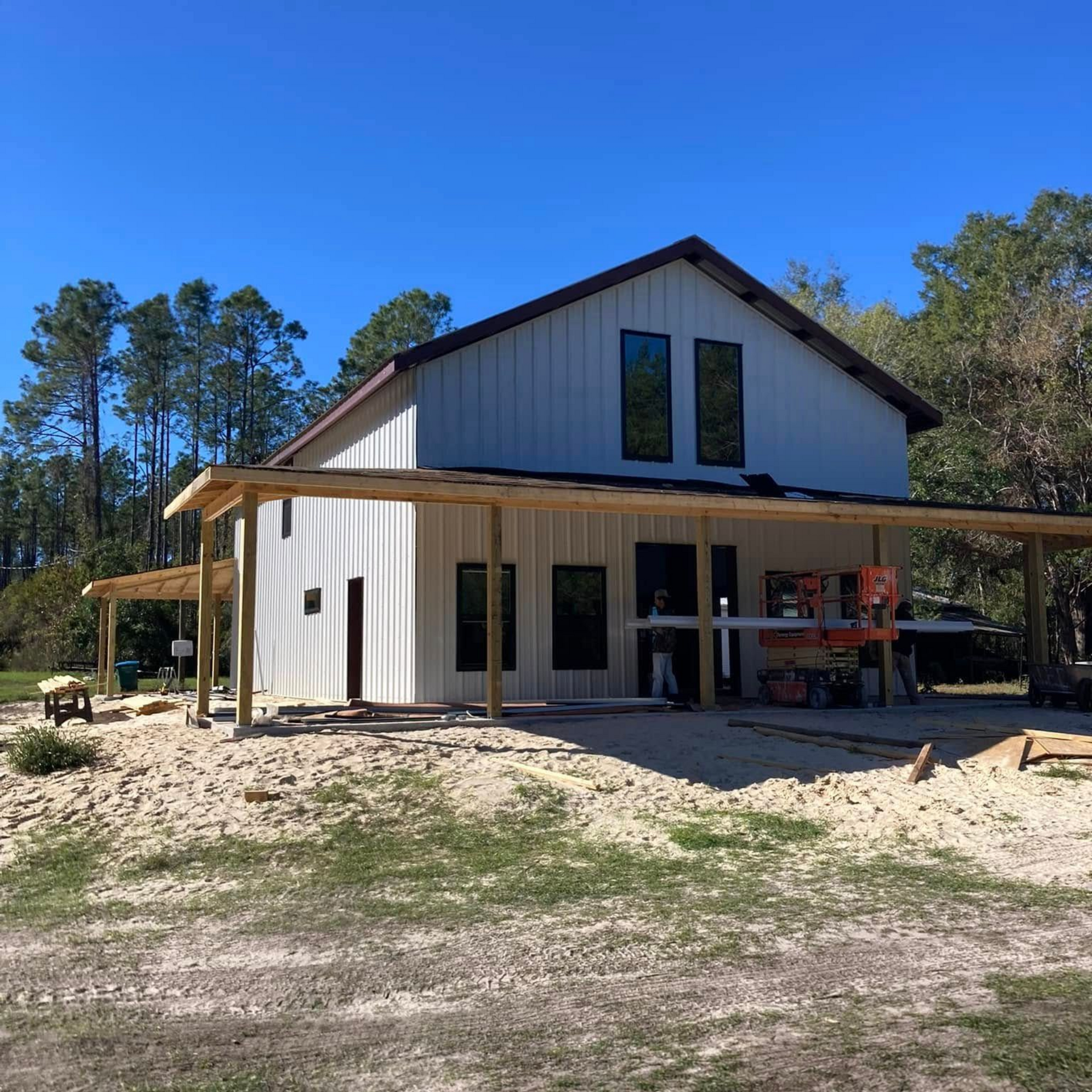 Two-story white house under construction with a covered porch and tall windows, surrounded by pine trees and dirt ground