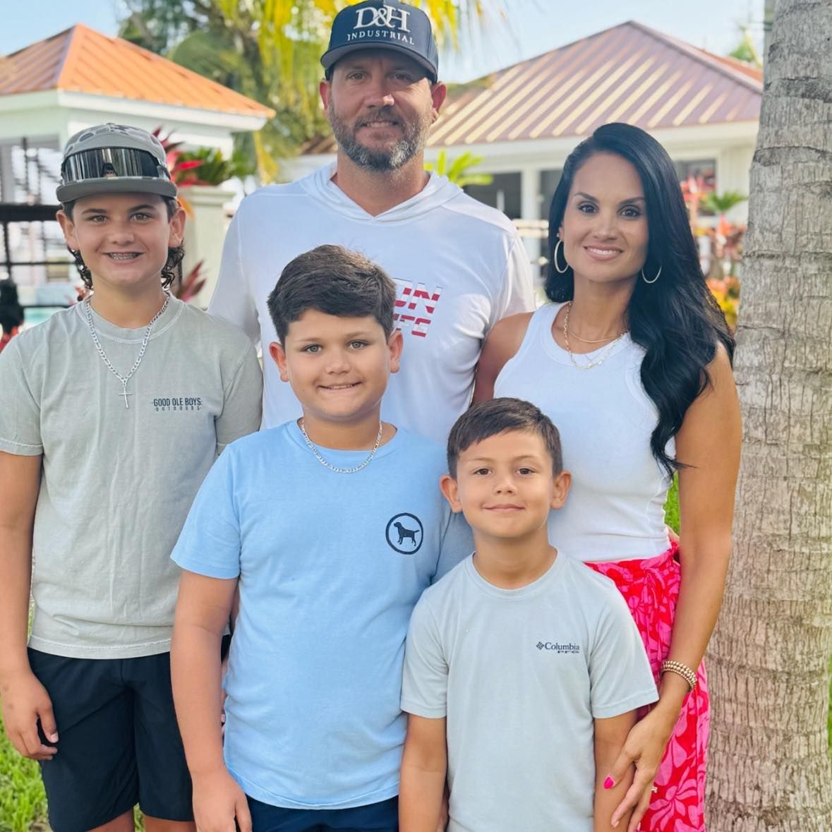 Family of five posing outdoors by palm trees, with a house in the background.
