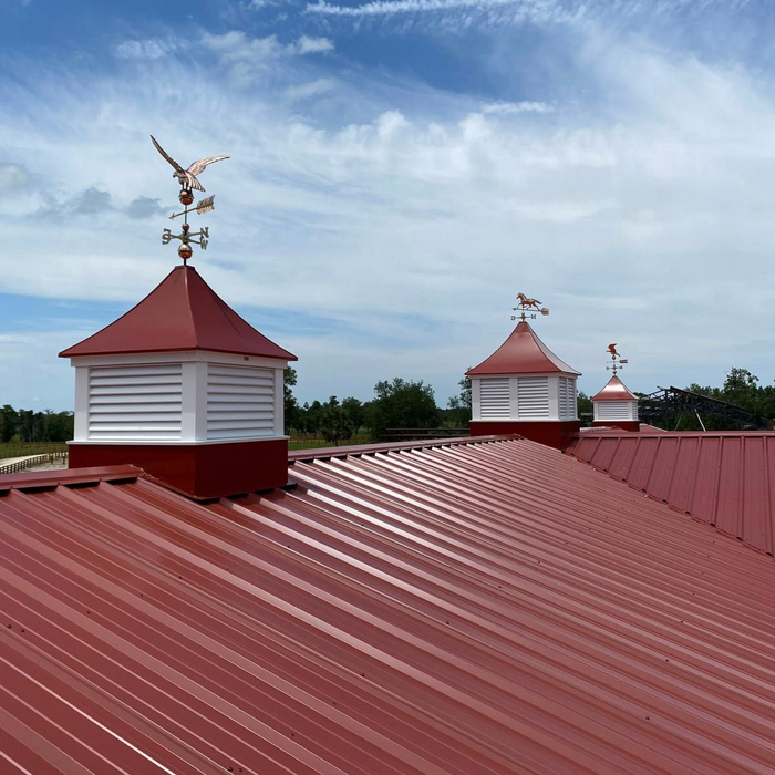 Red metal barn roof with white cupolas and weather vanes under a partly cloudy sky