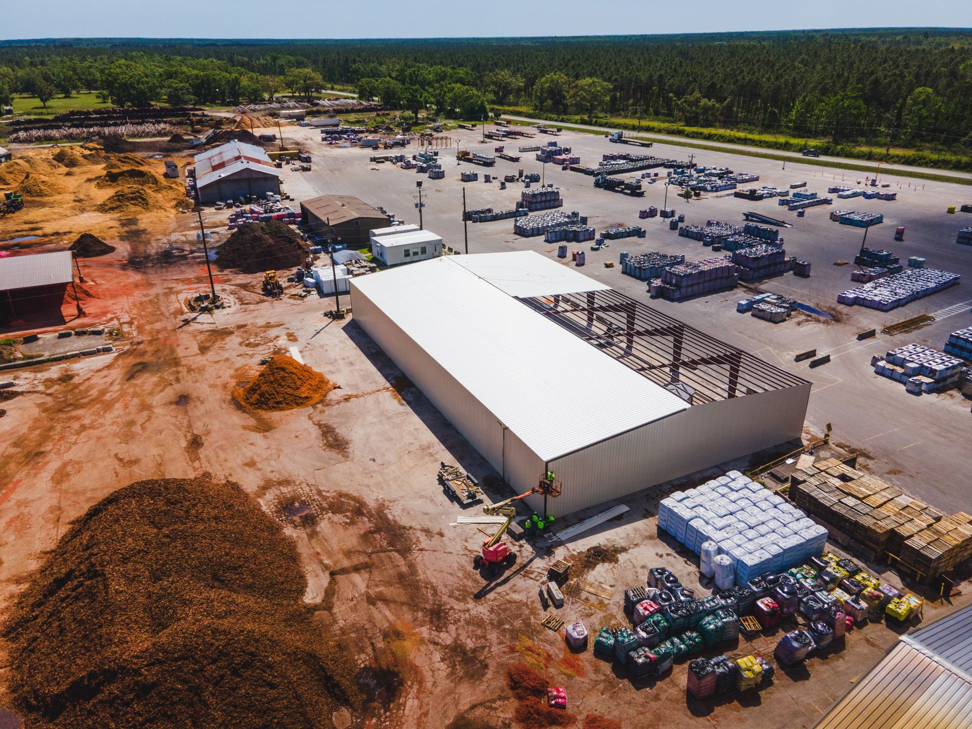 Aerial view of a warehouse yard with trucks, parked cars, and stacked materials beside a wooded area