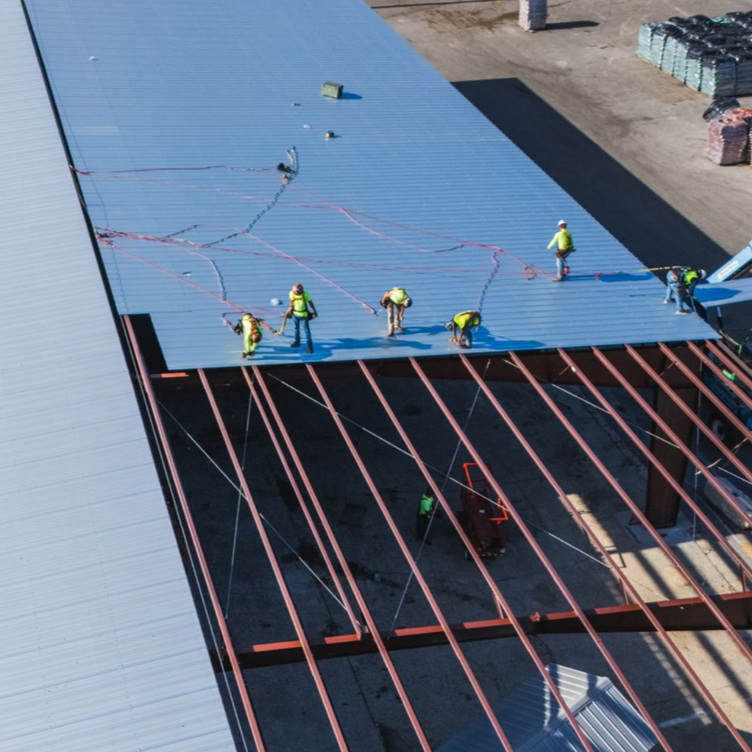 Workers installing a blue roof panel on a building under construction, viewed from above.