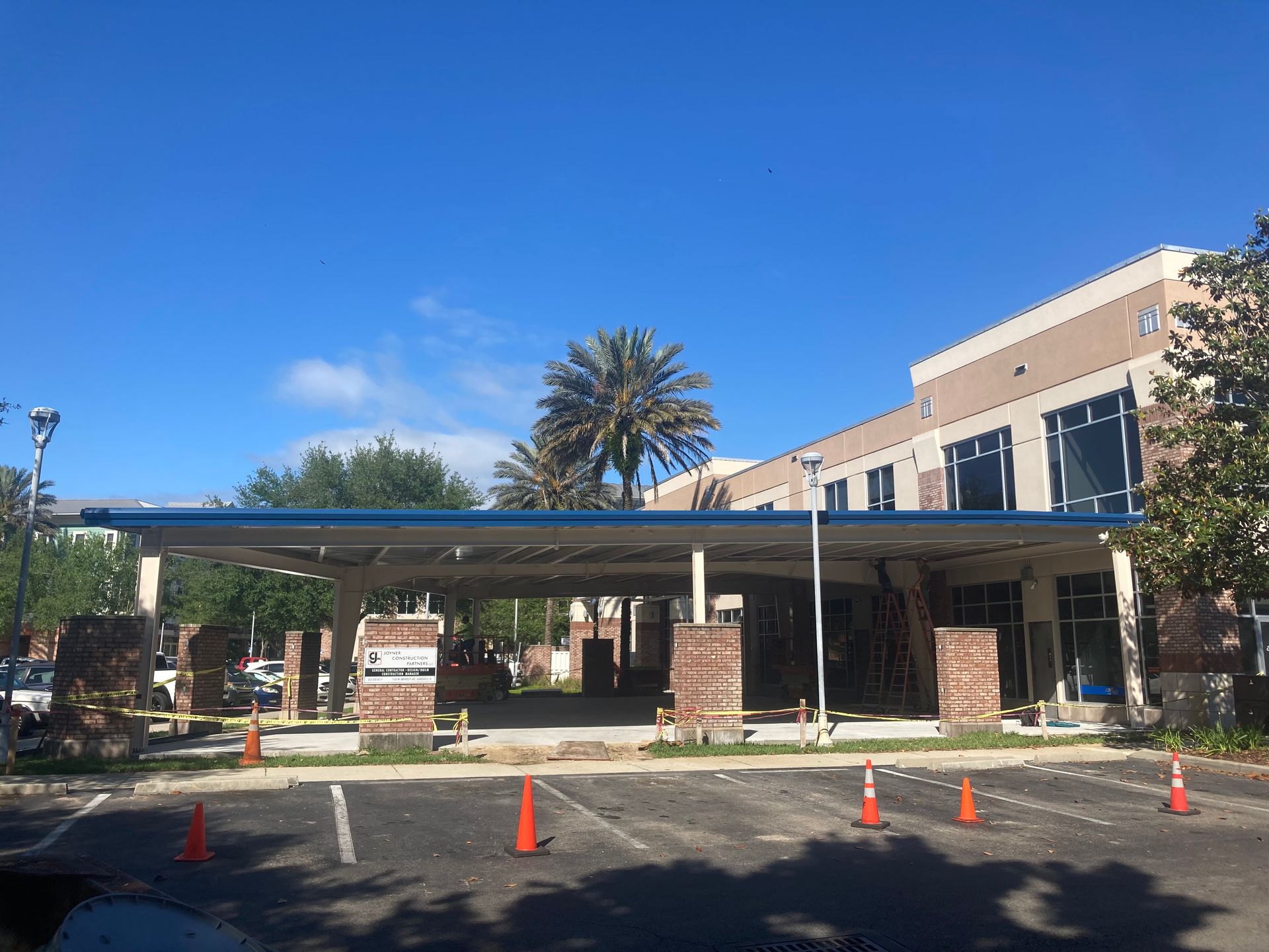 Building entrance with covered drop-off area, brick columns, palm trees, and orange traffic cones under a blue sky