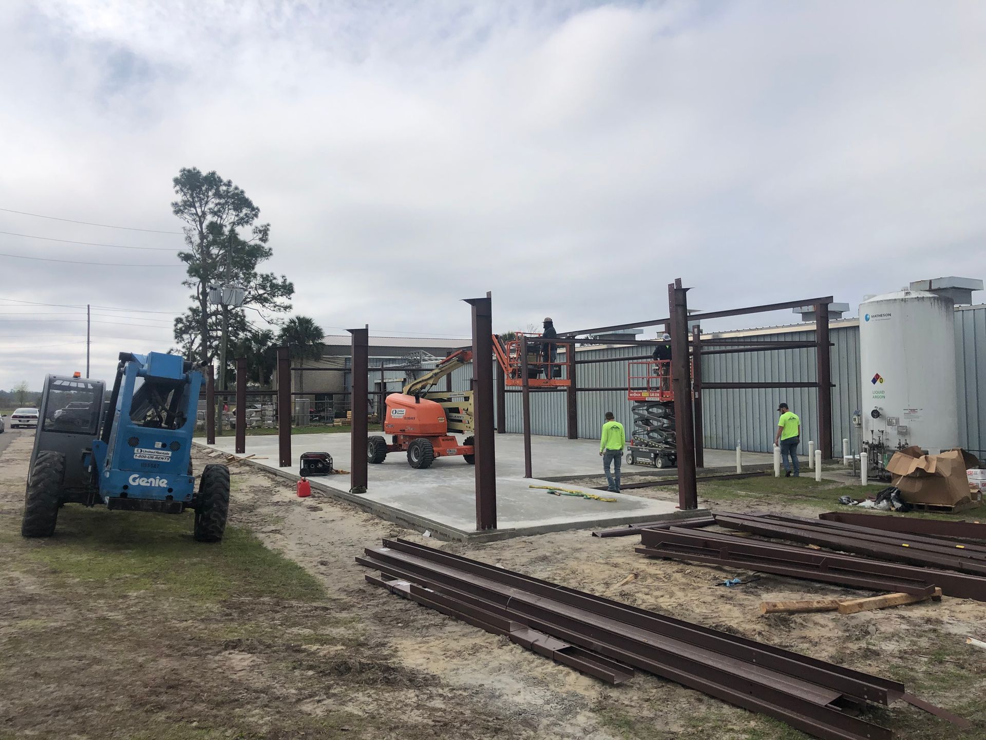 Construction site with steel frame, workers, orange lift, and blue forklift beside railroad tracks