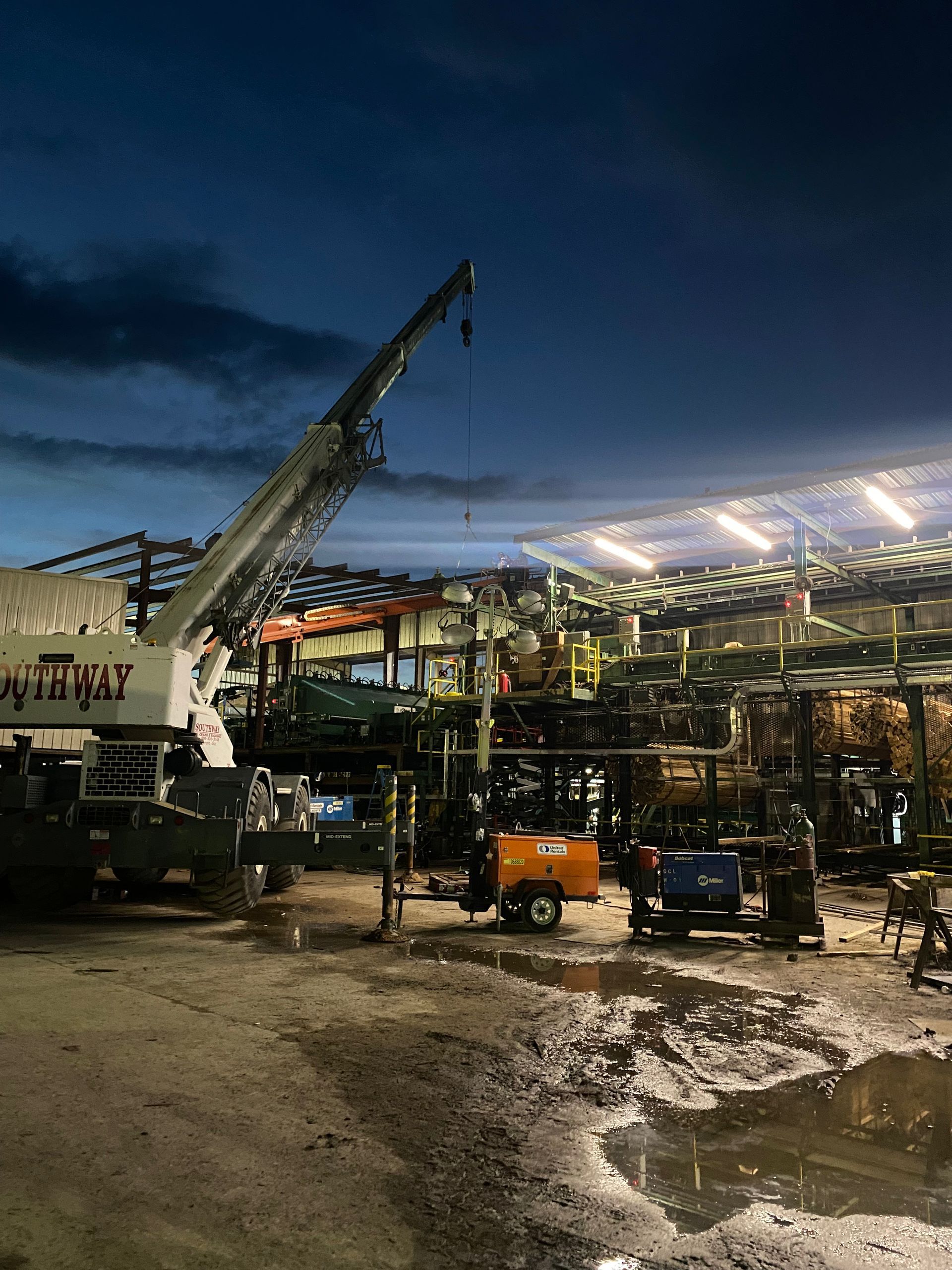 Nighttime construction site with a crane, steel framework, and bright lights reflecting in muddy ground.