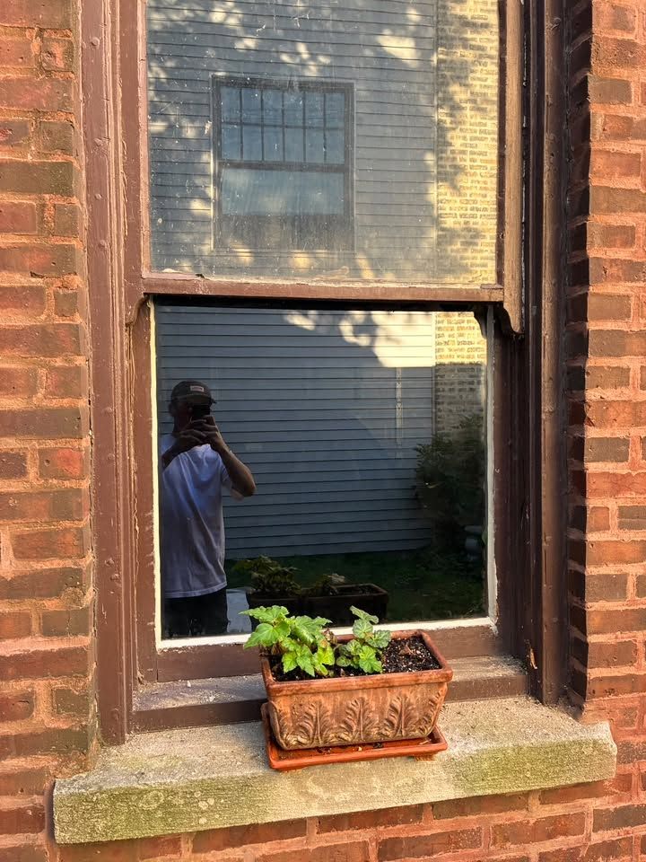 Window with a flower box; reflection shows a person taking a photo outside a brick house.