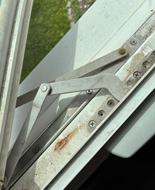 Close-up of a metal window hinge with rust spots on a white frame.