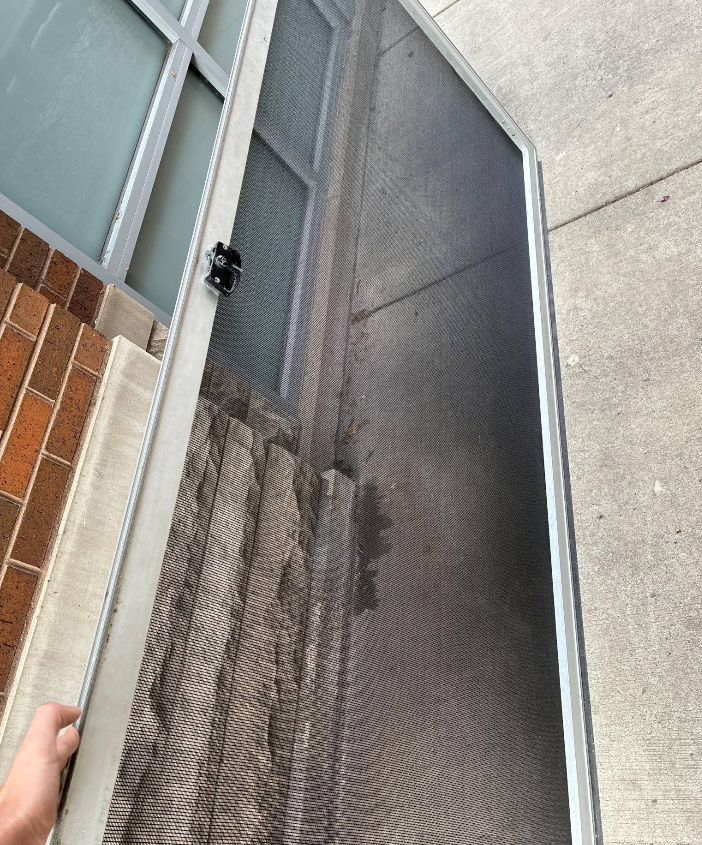 Close-up of a damaged mesh screen door beside a brick wall and concrete walkway