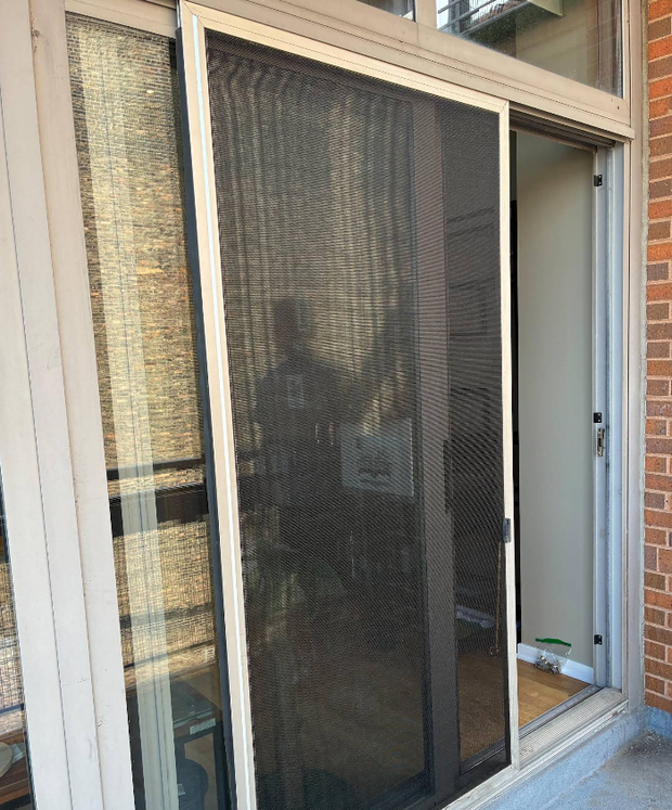 Open sliding screen door on a screened porch, with sunlight and a brick wall beside it.