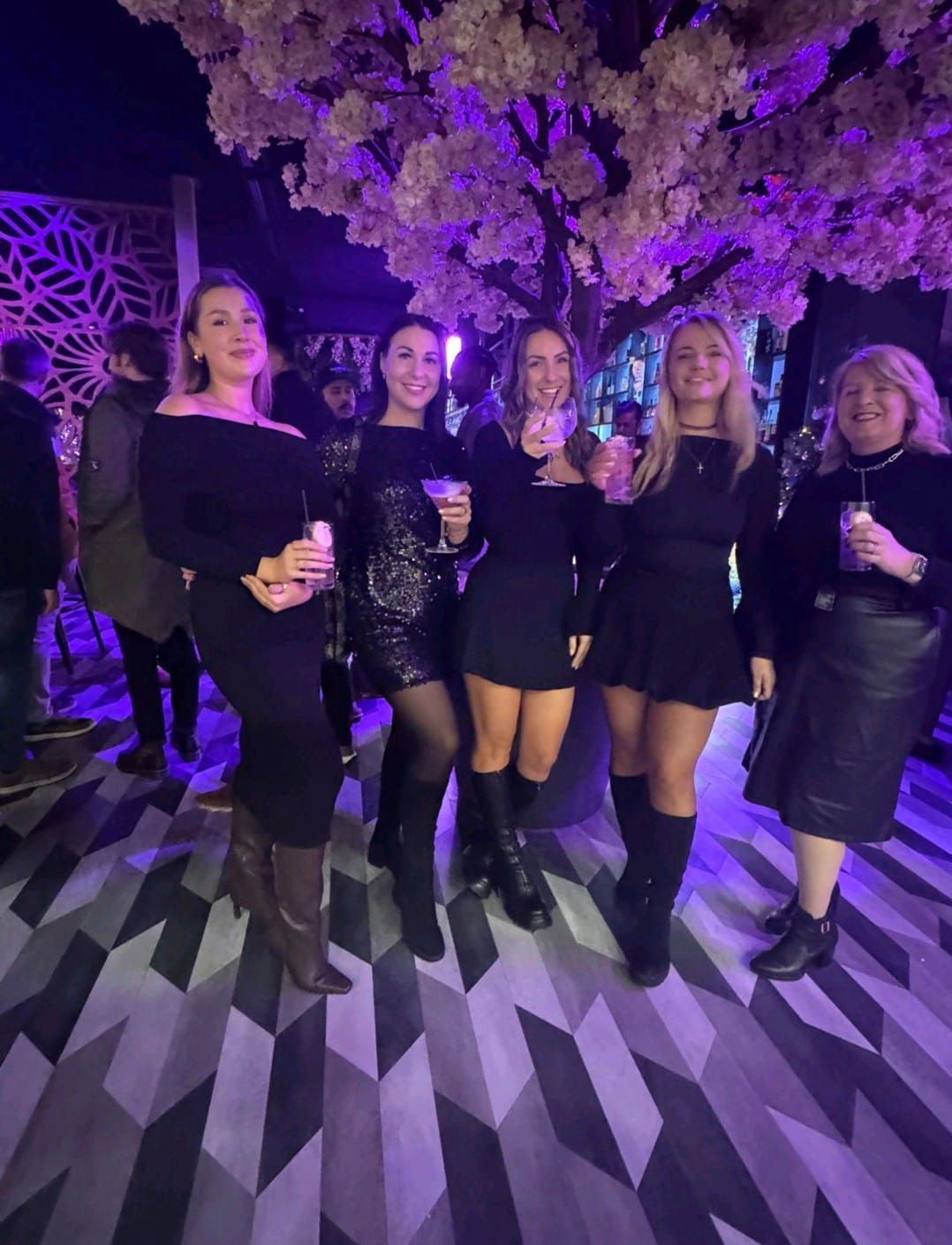 Five people holding drinks pose in black outfits at a purple-lit party.