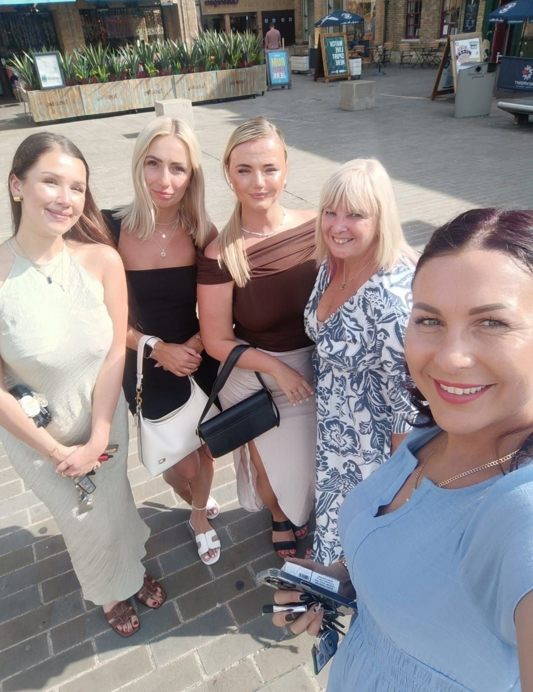 Five women smiling for a selfie outdoors on a sunny day