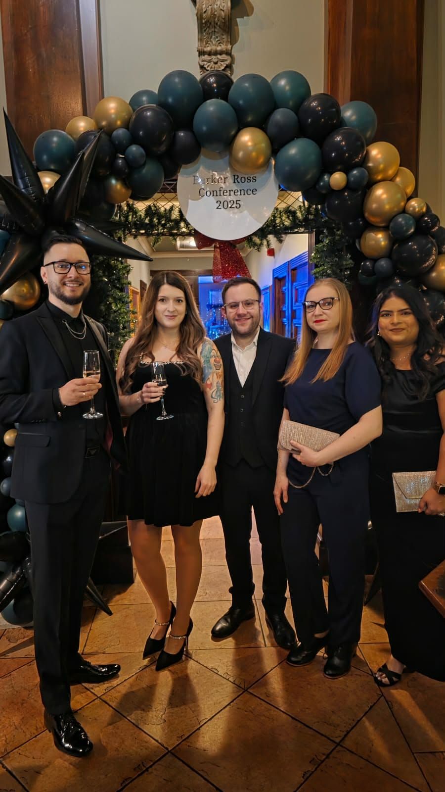 Five people posing under a black and gold balloon arch at an indoor event