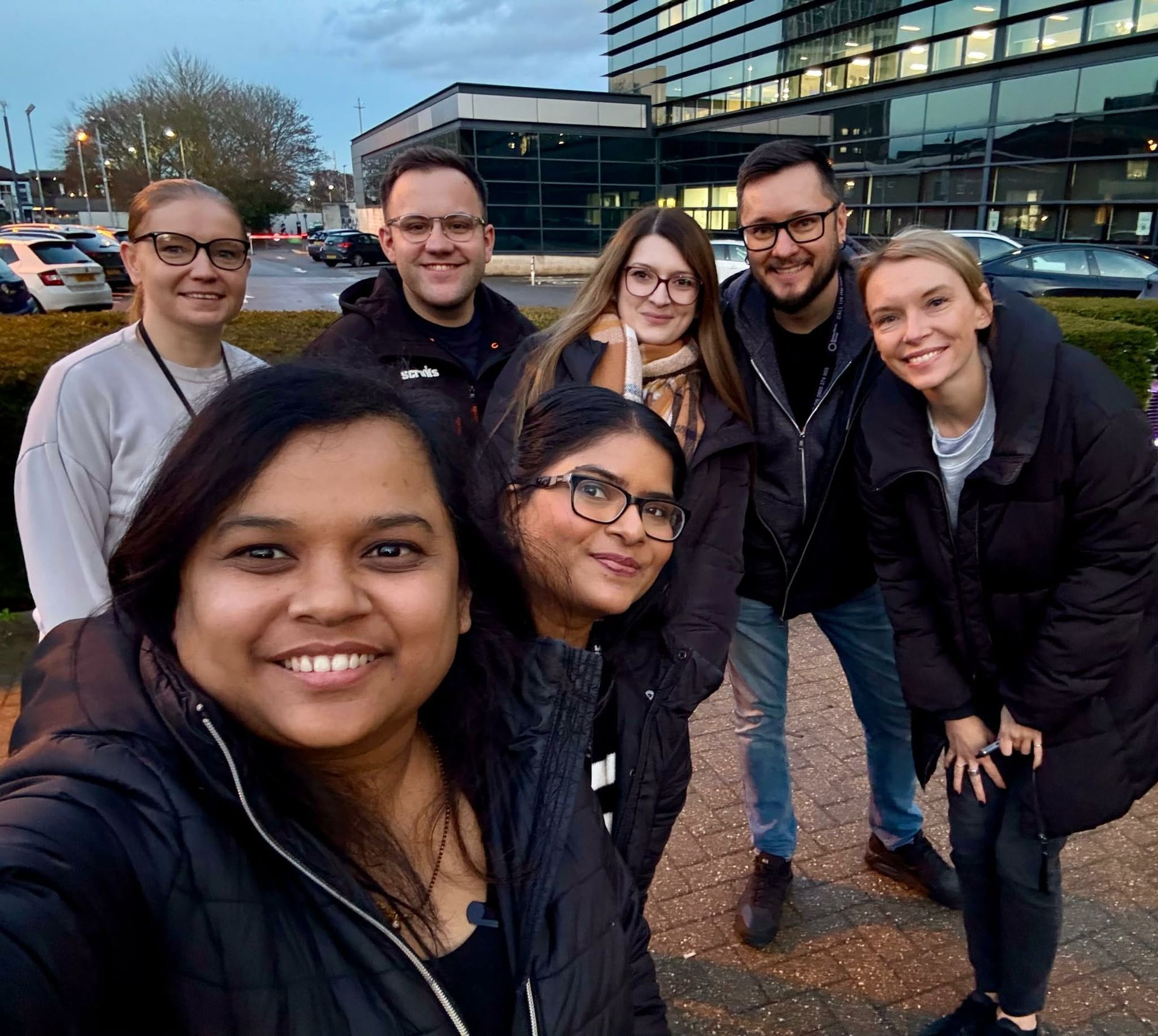 Group of six smiling coworkers posing outdoors beside a modern glass office building at dusk