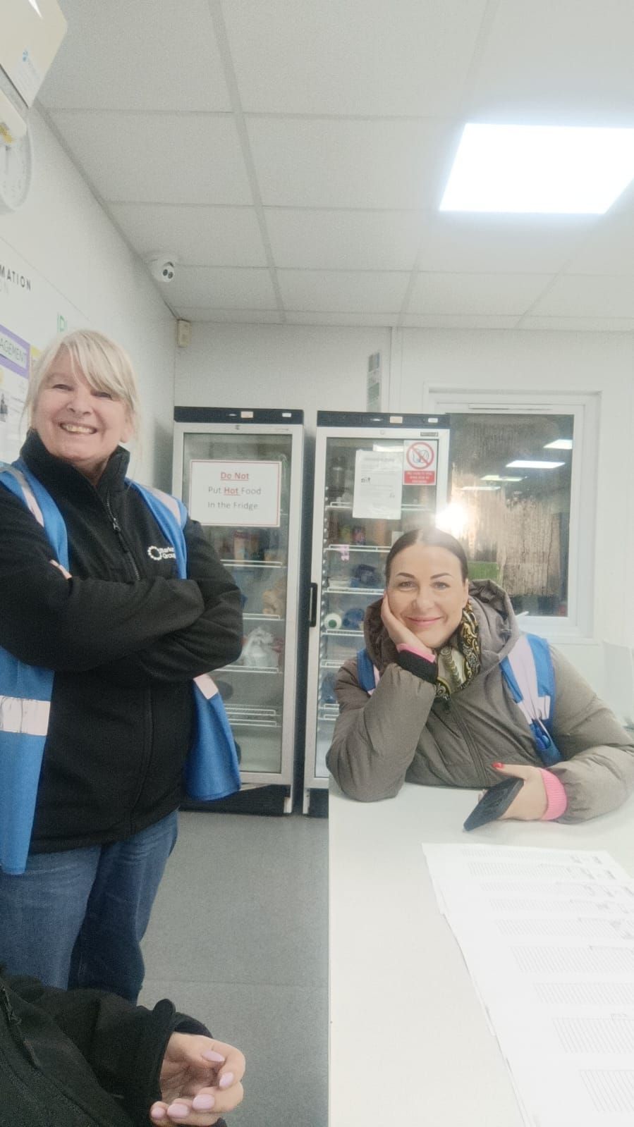 Two people smiling in an office break room, one standing by a fridge and one seated at a table.