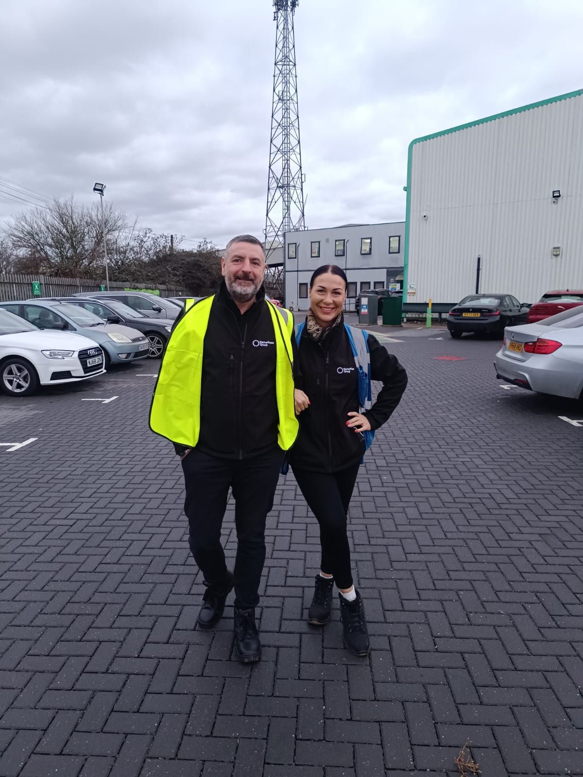 Two people standing in a parking lot, one in a neon yellow jacket, smiling at the camera