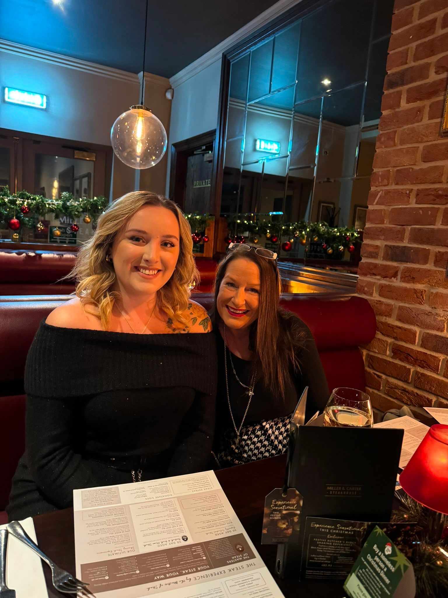 Two smiling women seated at a restaurant table with menus, in front of a large window at night
