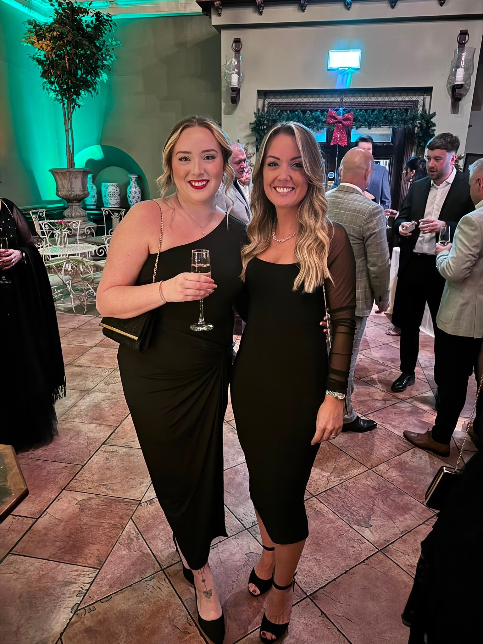 Two women in black dresses holding drinks and smiling at a formal indoor event.