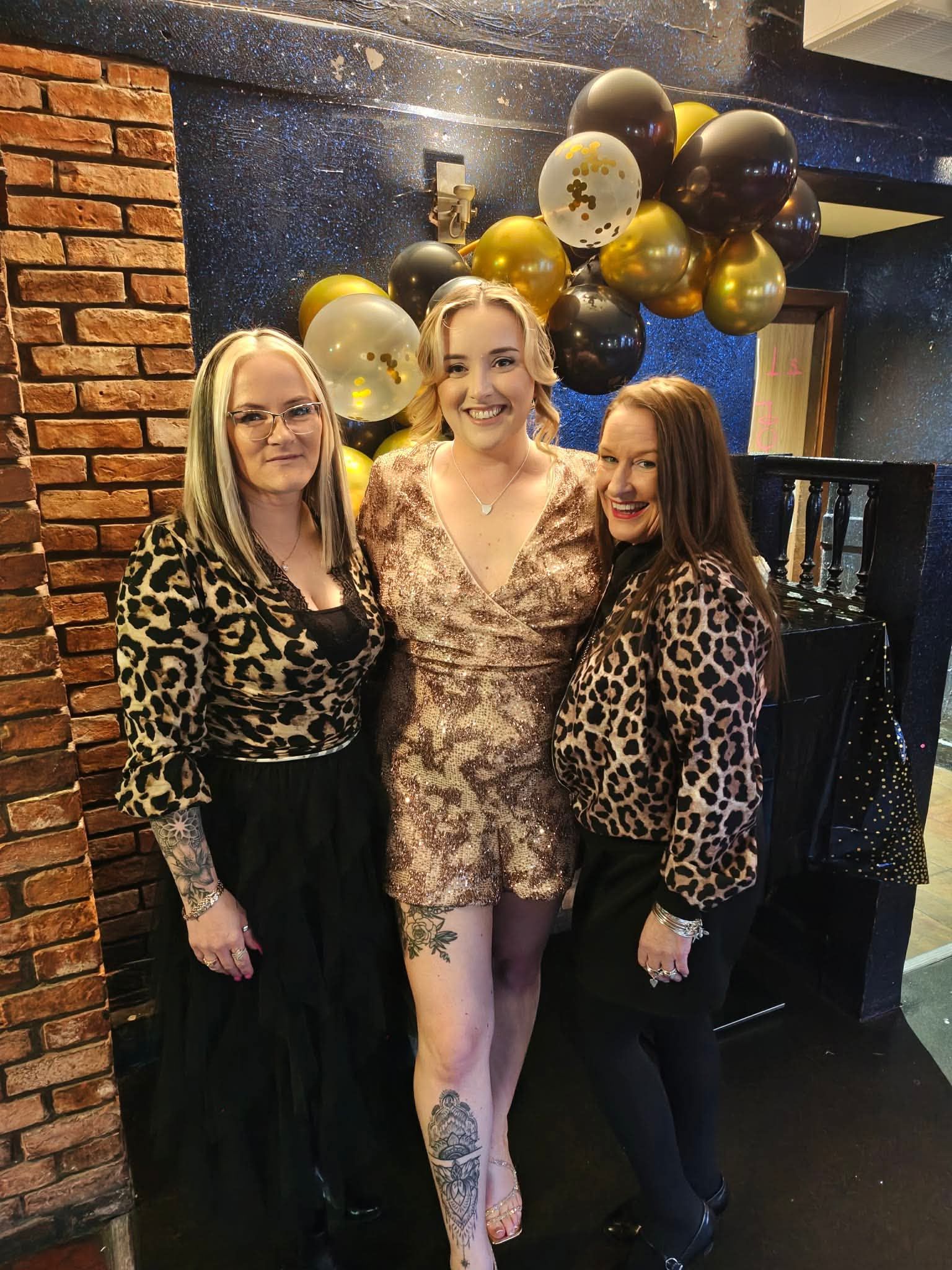 Three women posing at a party with gold and black balloons, wearing leopard print outfits.