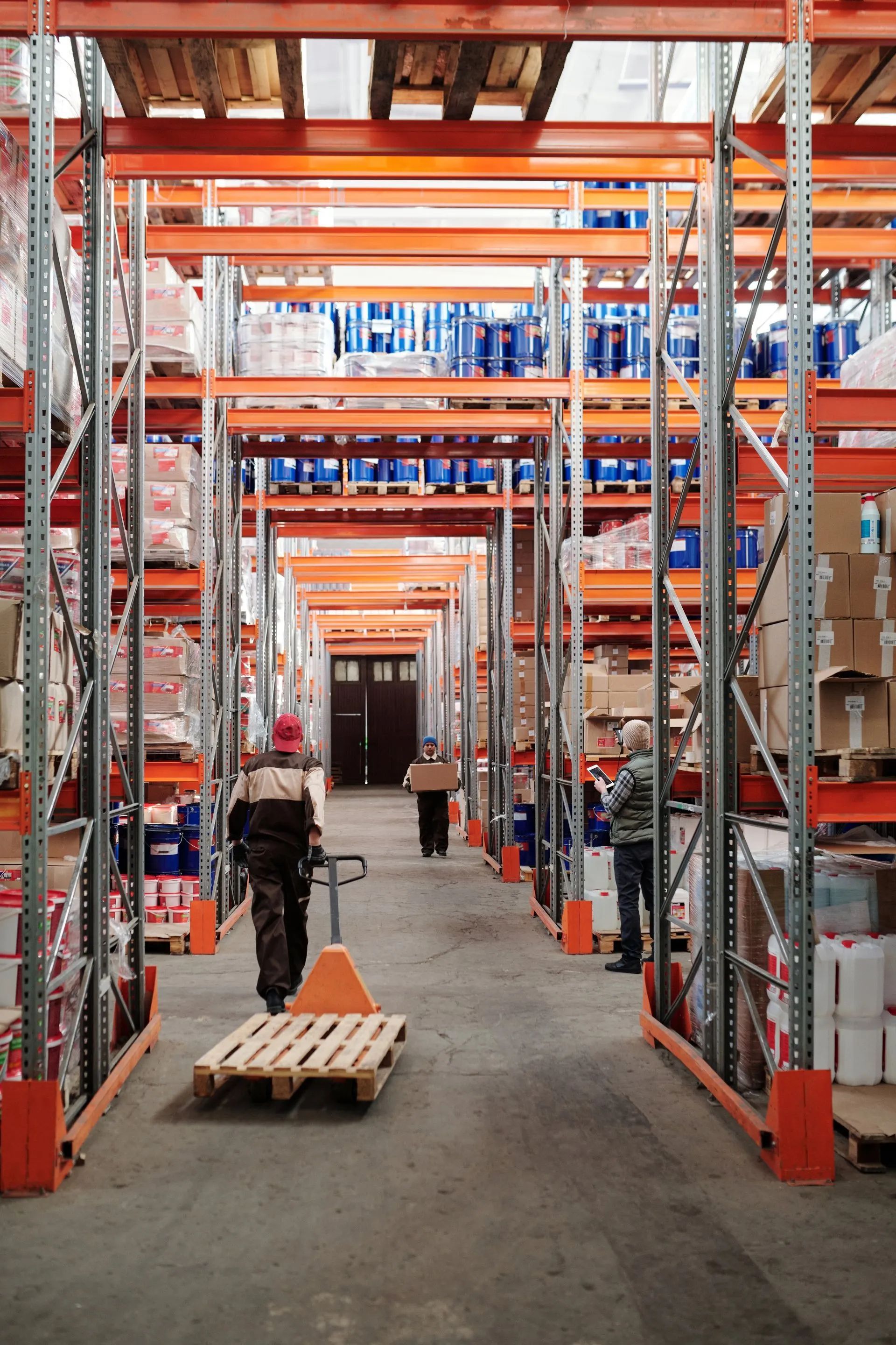 Warehouse aisle with tall orange racks, stacked boxes, and a worker moving a pallet jack down the aisle