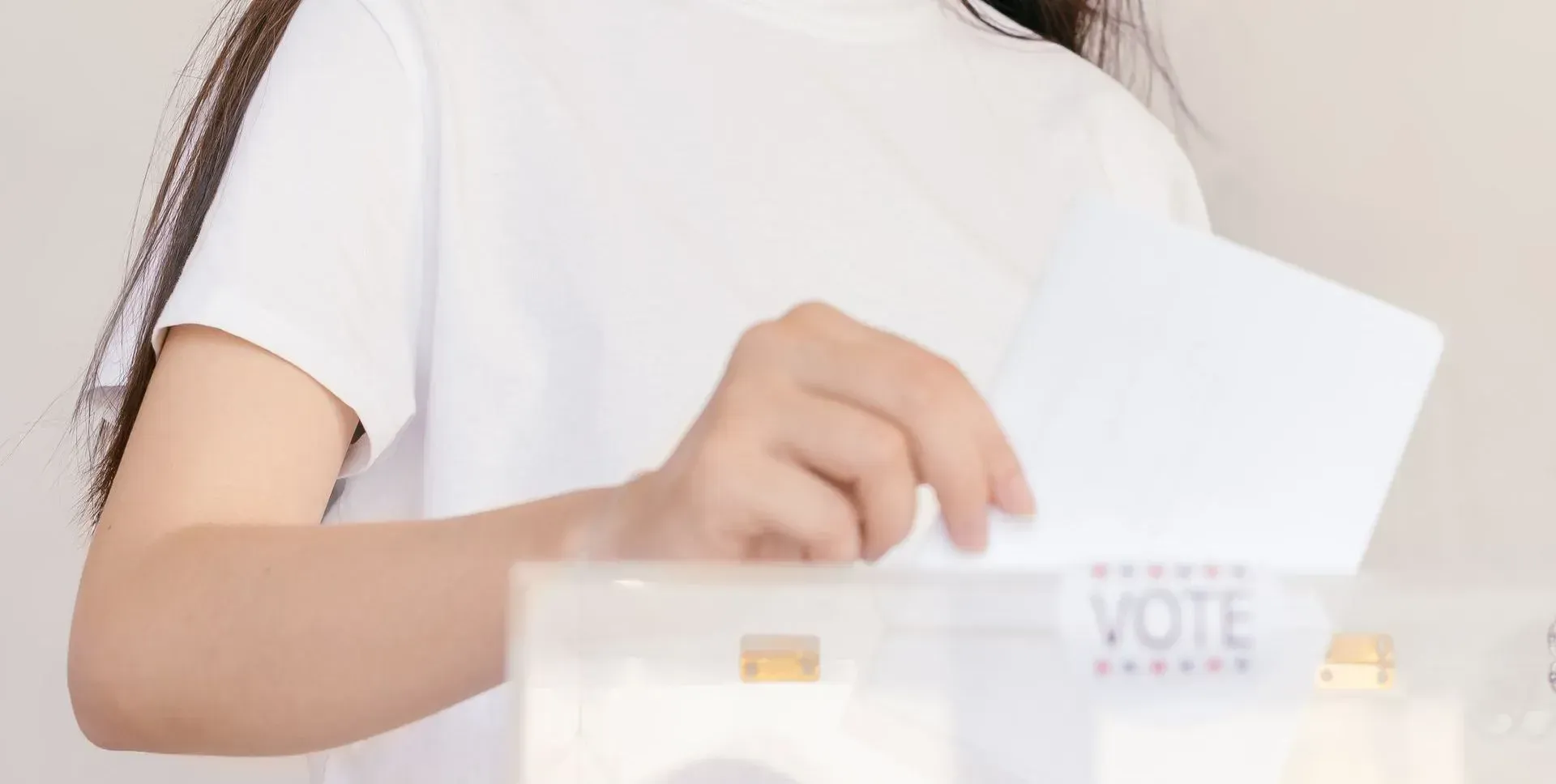 Hand placing a ballot into a transparent election box labeled “VOTE”