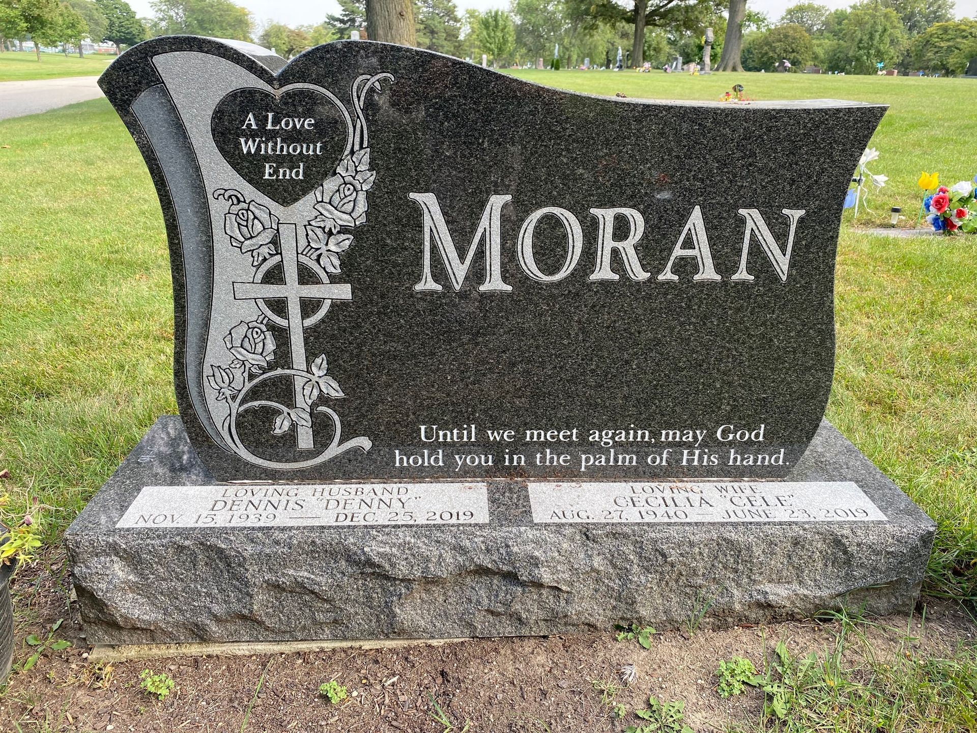 A black granite headstone with the name MORAN and an engraved cross and heart, set in a green grassy cemetery.