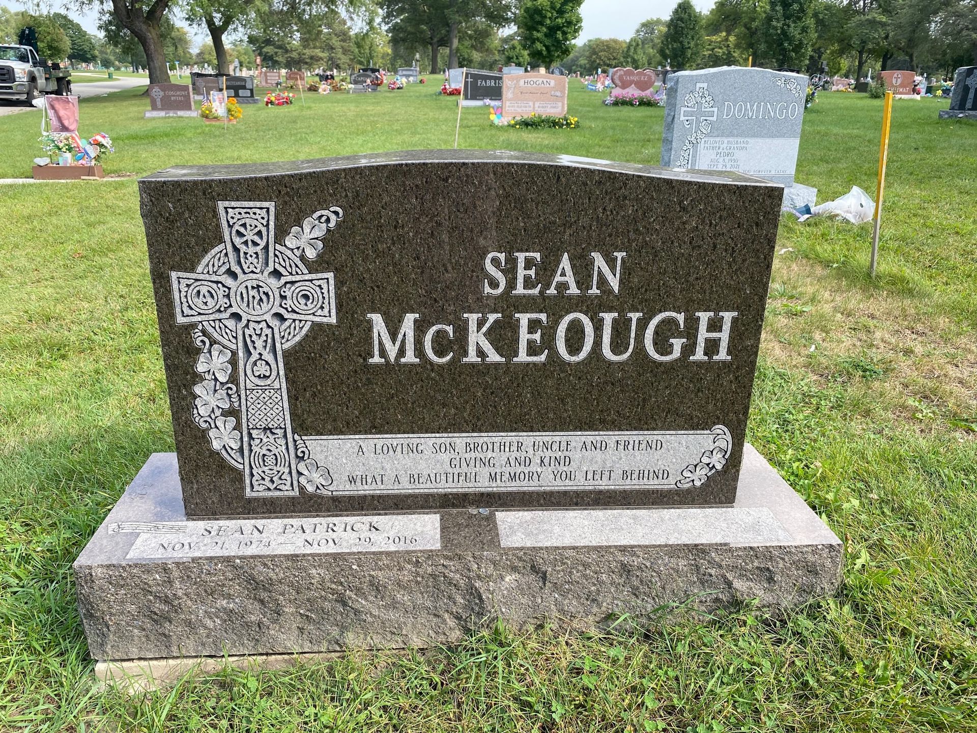 A dark granite headstone for Sean McKeough, inscribed with a Celtic cross, set in a sunny, green cemetery.