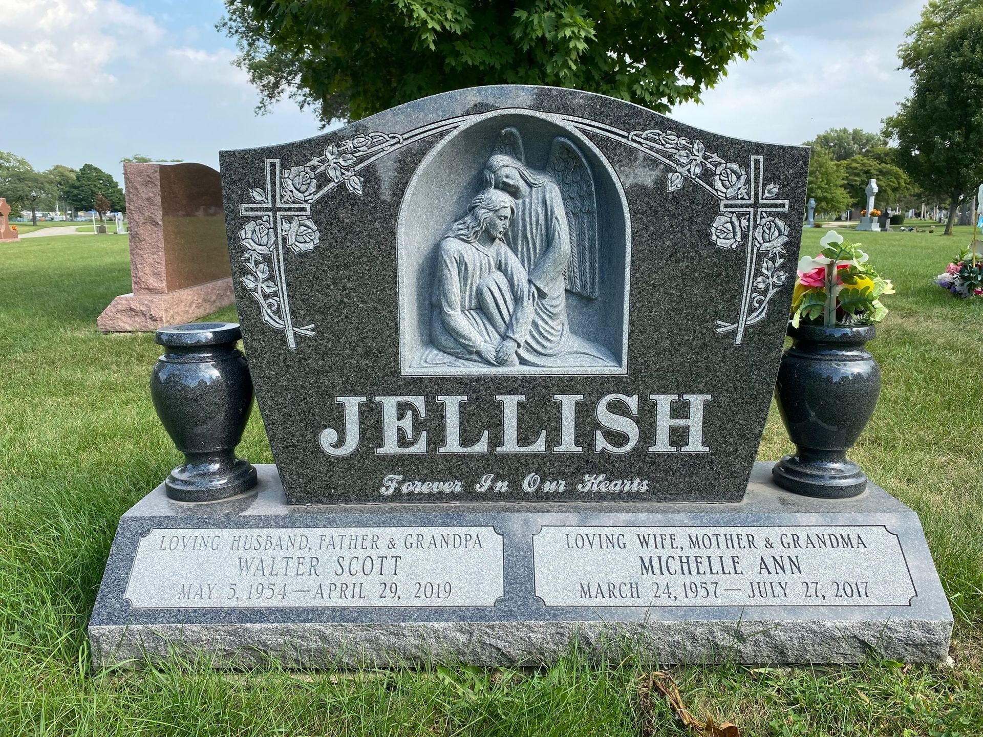 A gray granite Jellish family headstone in a cemetery, featuring a carved angel and two plaques with memorial dates.