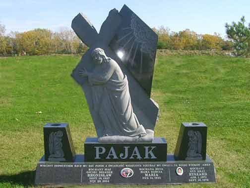 A granite tombstone featuring a carved statue of Jesus carrying a cross, set in a grassy cemetery.
