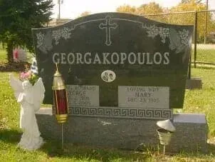 A black granite Georgakopoulos family headstone with a cross, memorial lamp, and angel statue in a grassy cemetery.