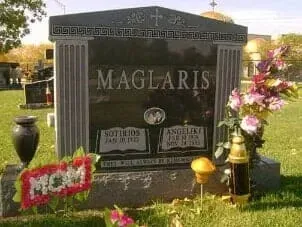 A black upright monument for the Maglaris family in a cemetery, with floral tributes and a small gold lamp at its base.