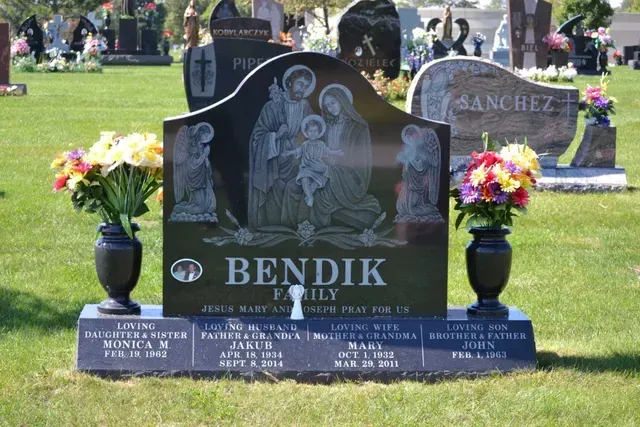 Black polished granite headstone for the Bendik family, featuring an etching of the Holy Family, with floral arrangements.