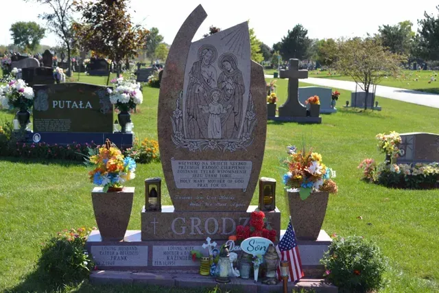 A tall, reddish-brown granite tombstone for the Grodzki family, featuring an etched holy family, in a sunny cemetery.
